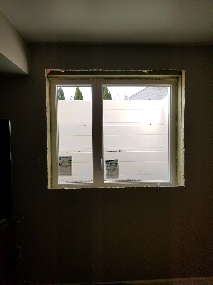 A window installed in a dark room with unpainted drywall edges and white protective film still on the glass panes.