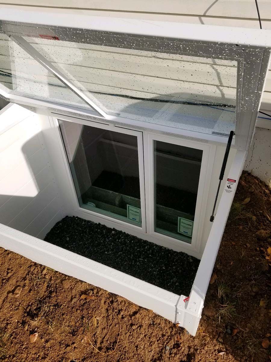 A white window well cover is propped open, revealing a white basement window set in a gravel-filled enclosure.