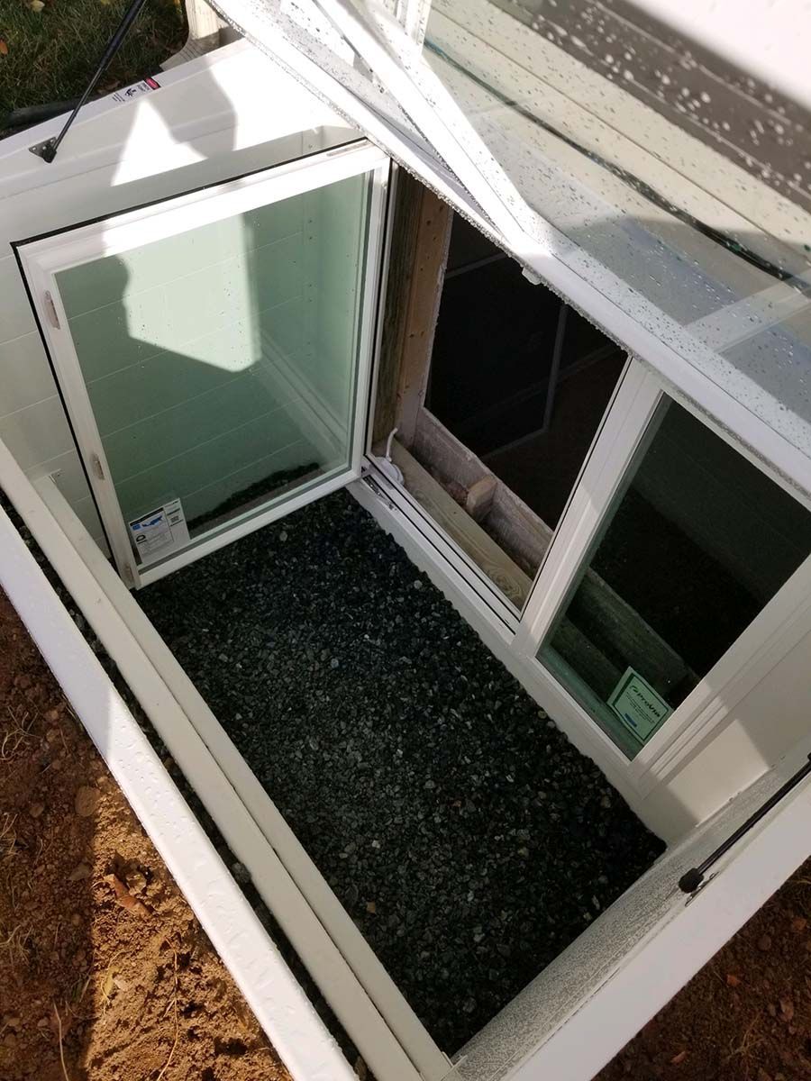 A white frame surrounding a basement window well with a black mesh cover and decorative stone inside.