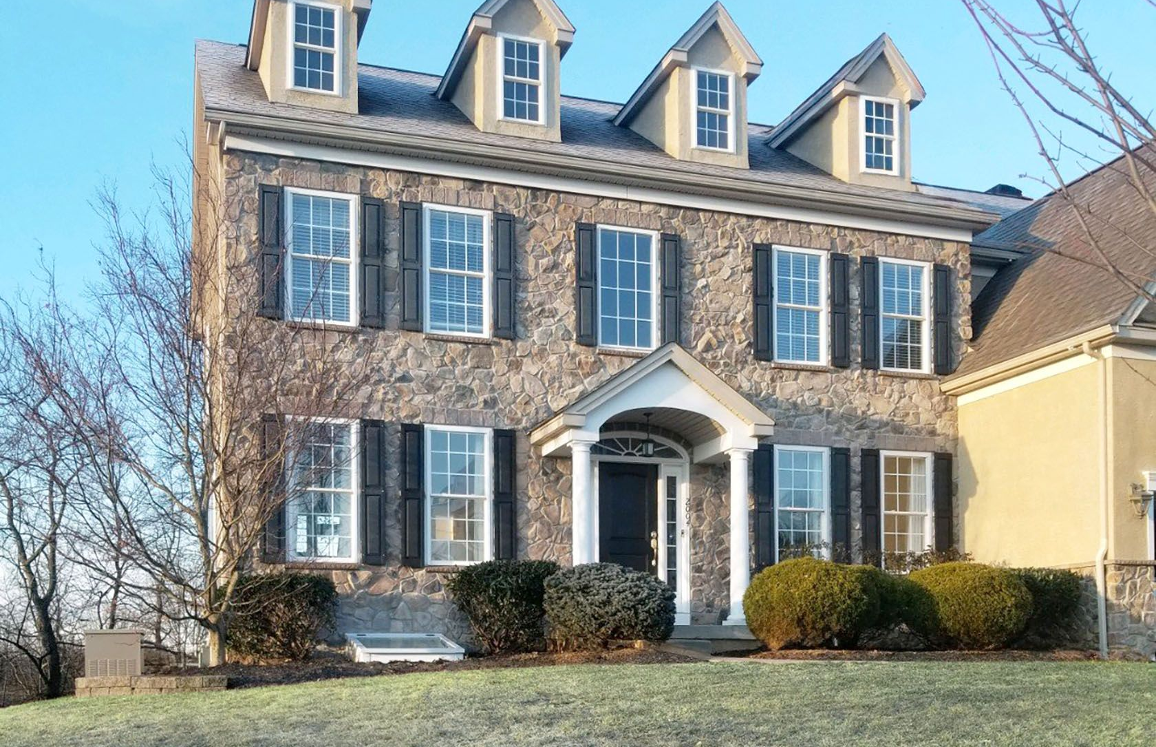 A two-story stone house with dark shutters, four dormer windows on the roof, and a front porch, set on a grassy hill.