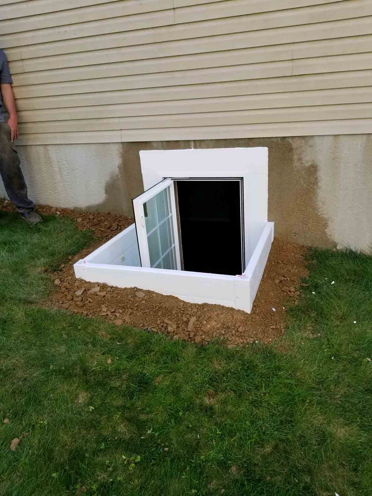 A white basement window well with an open window installed against a house foundation with exposed dirt around the base.