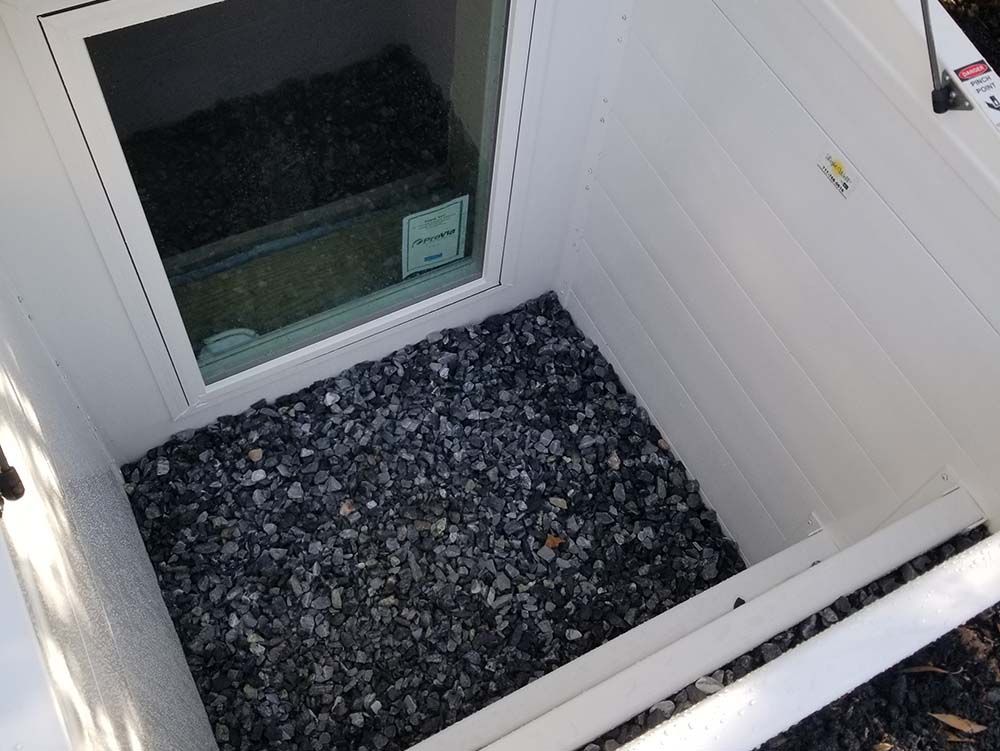 A white window well for a basement window, filled with dark grey crushed stone against a house foundation.