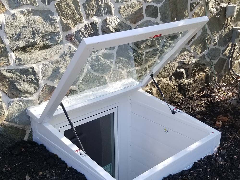 A white basement egress window with a transparent cover open, installed against a stone foundation wall.