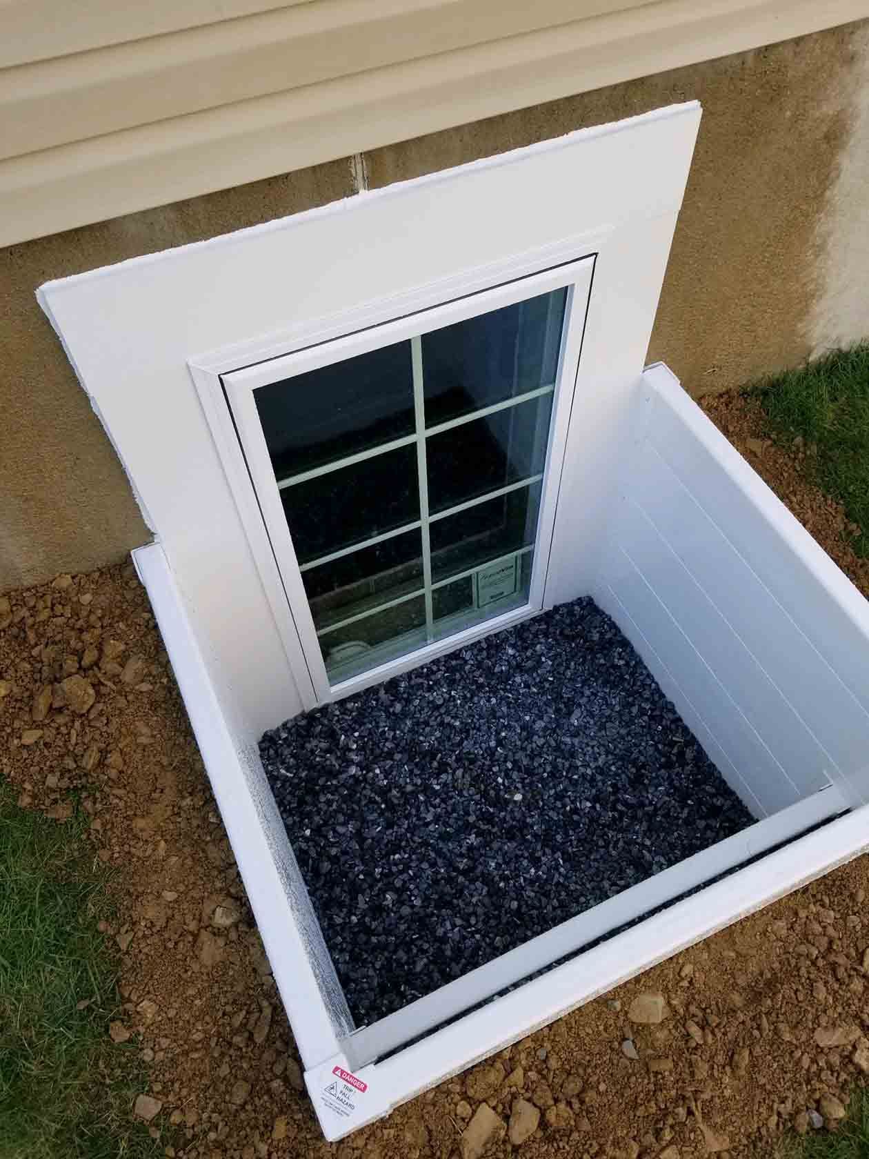 A white basement egress window with a matching white window well liner filled with black gravel, set in dirt and grass.