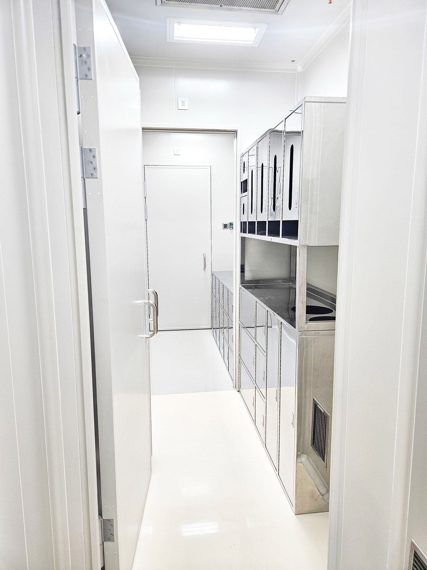 A bright, sterile, white-tiled narrow hallway leading to a door, with metal cabinetry lining the right wall.