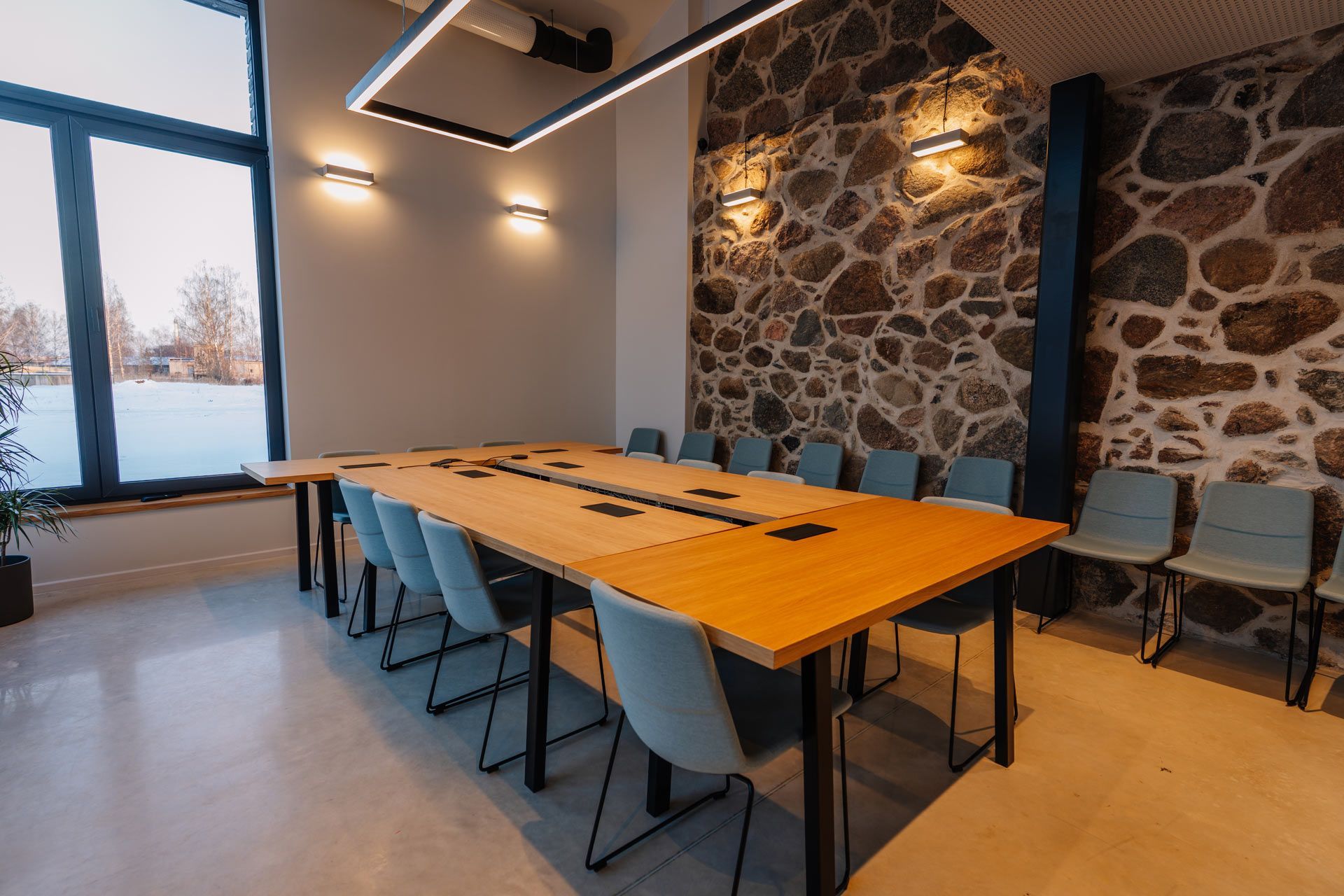 A long wooden conference table surrounded by light blue chairs in a room with a large window and stone feature wall.