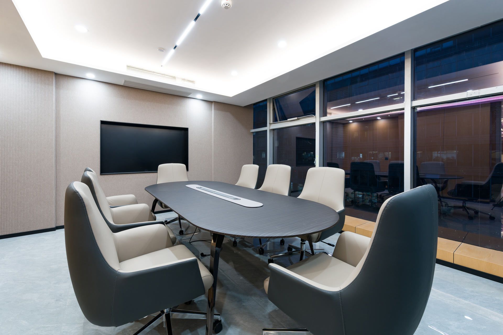 A modern, minimalist boardroom with a black oval table, gray chairs, light wood paneled walls, and large floor-to-ceiling windows.
