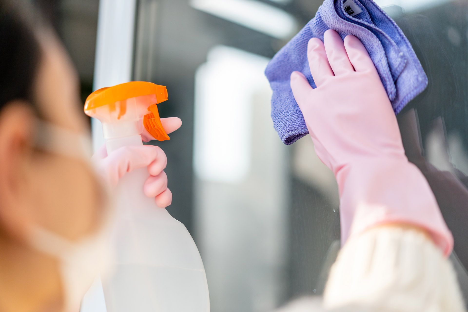 A person wearing pink gloves and a face mask wipes a glass surface with a cloth while holding a spray bottle.