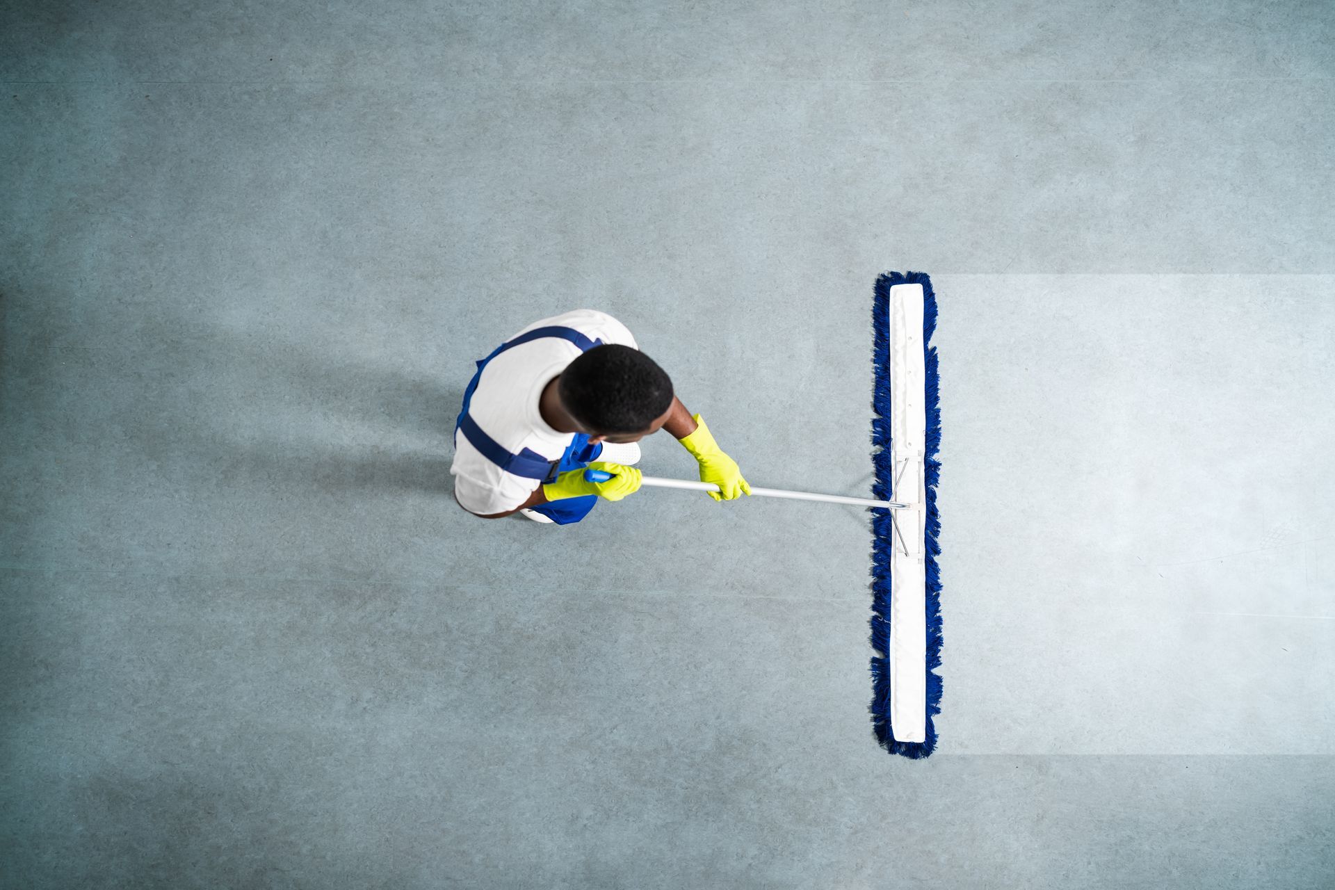 Overhead view of a person using a wide mop to clean a smooth gray floor surface.