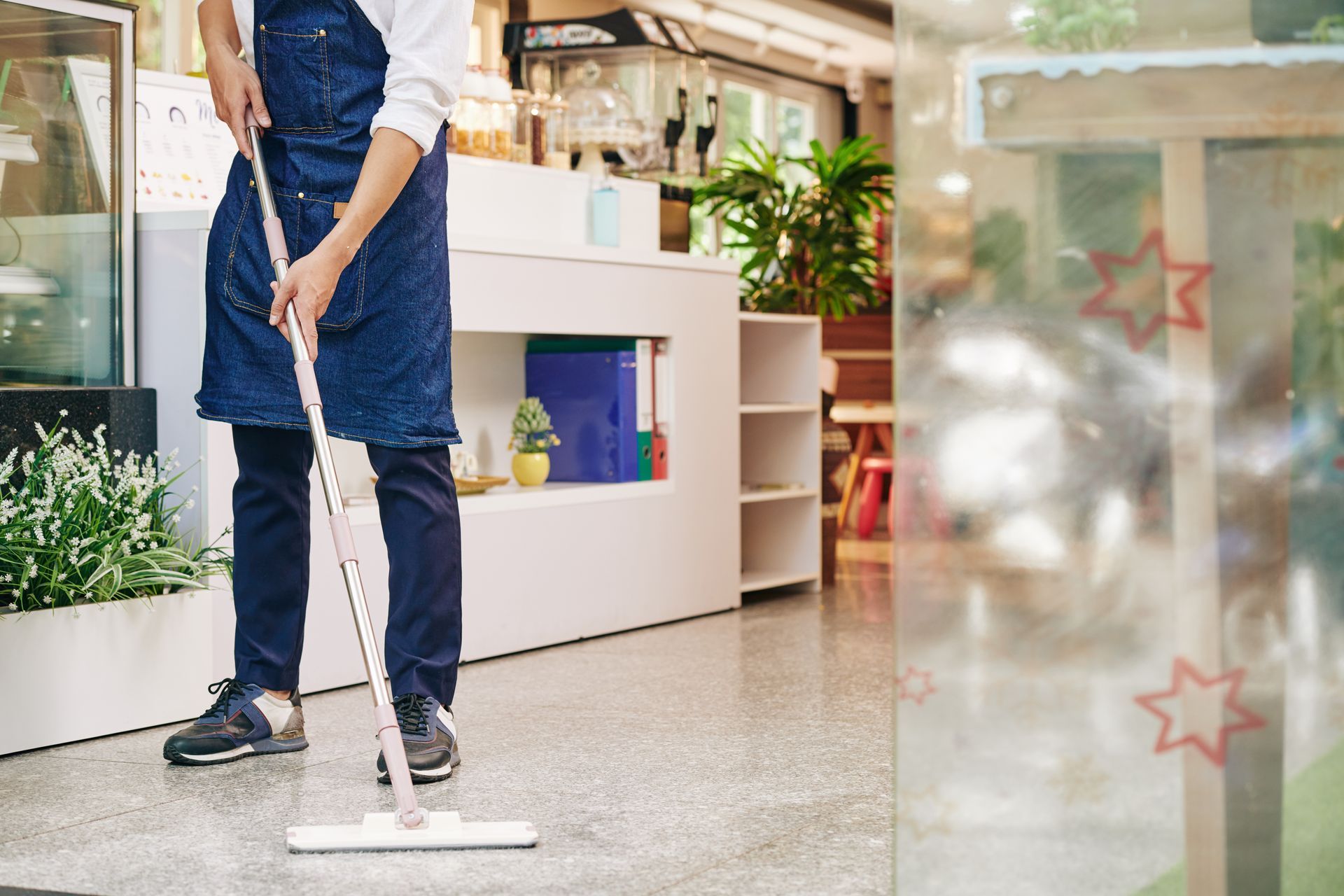 Person mopping a gray floor inside a cafe, wearing an apron. Person mopping a gray floor inside a cafe, wearing an apron.