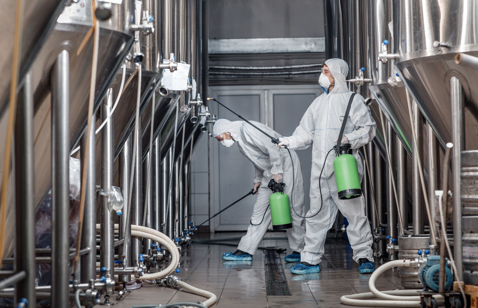 Two workers in protective suits spraying disinfectant in a brewery, among metal tanks and pipes. Two workers in protective suits spraying disinfectant in a brewery, among metal tanks and pipes.