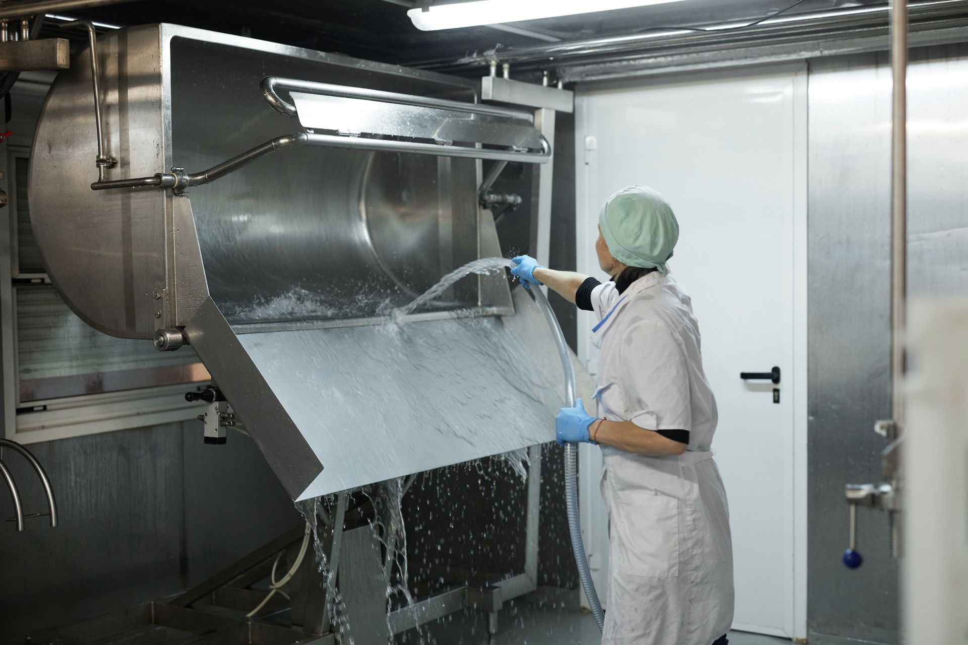 A person in a hairnet and lab coat sprays water onto a metal surface in a food processing setting. A person in a hairnet and lab coat sprays water onto a metal surface in a food processing setting.