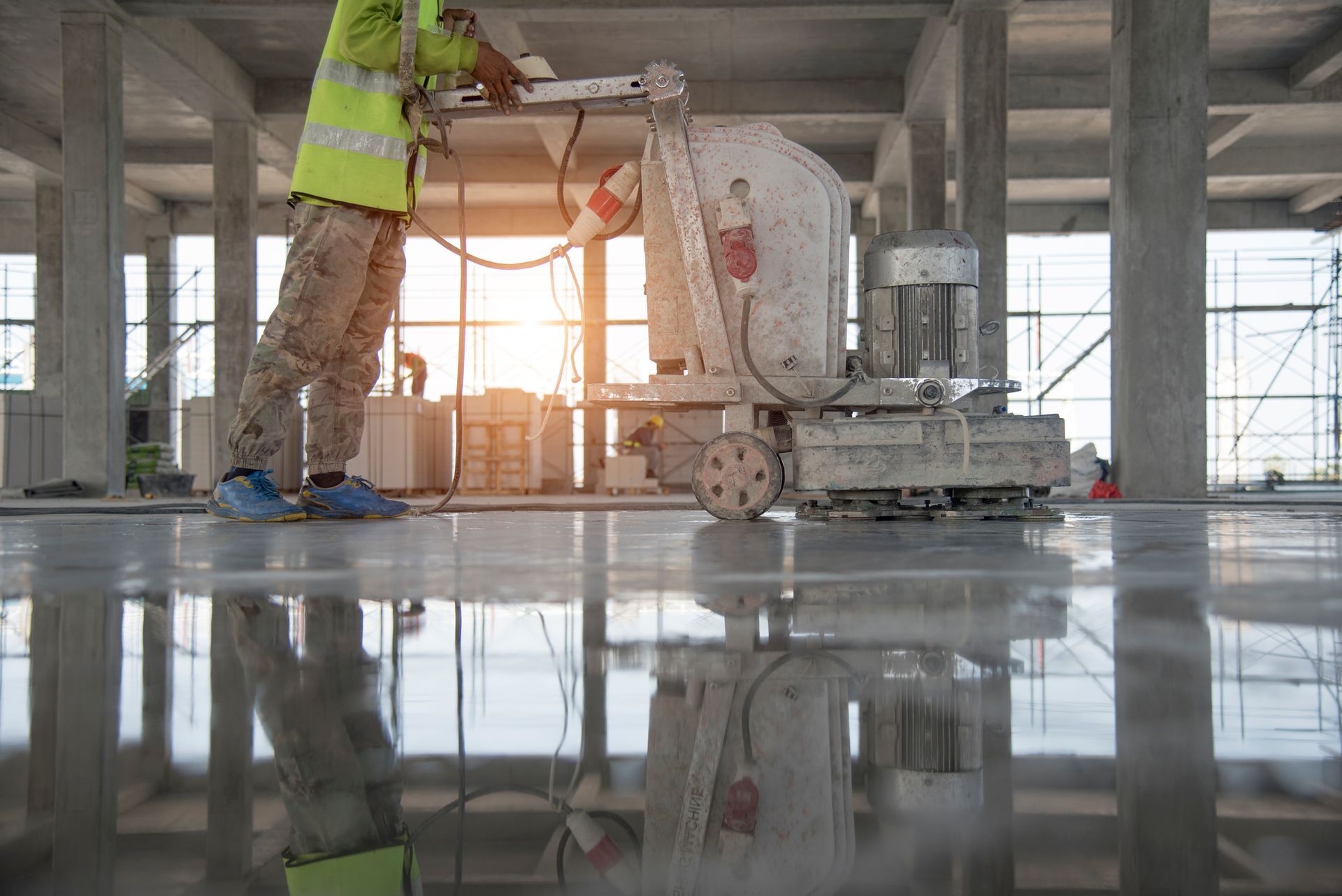 Person using a floor grinder on a reflective concrete surface in a construction site.