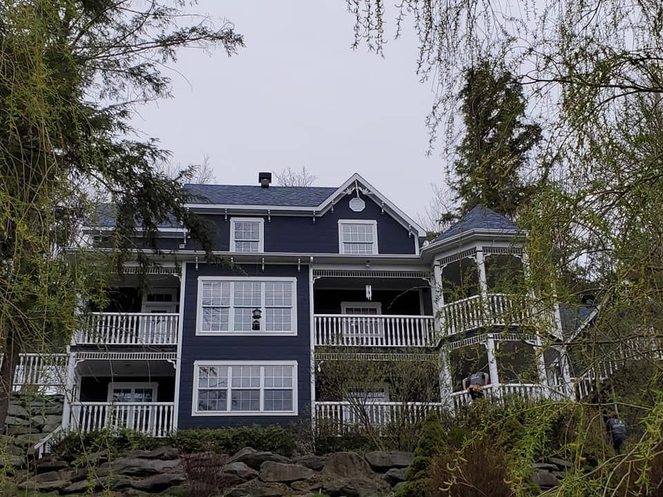Maison bleue à deux étages avec boiseries blanches, porche et kiosque, vue cachée derrière les arbres. Ciel nuageux.