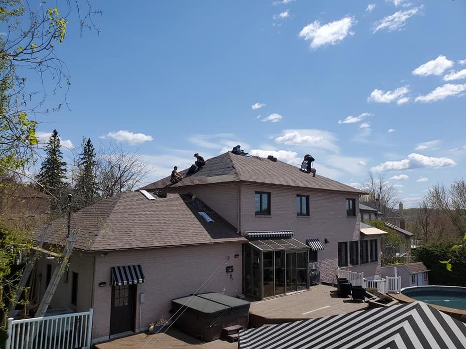 Des ouvriers posent des bardeaux sur le toit d'une maison à deux étages sous un ciel bleu parsemé de nuages.