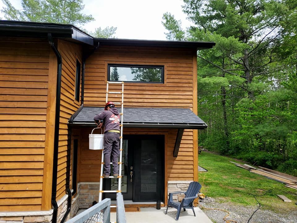 Une personne, perchée sur une échelle, peint l'extérieur d'une maison en bois à deux étages. Pelouse verte en arrière-plan.