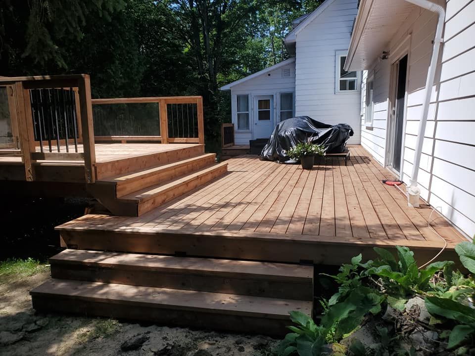 Terrasse en bois avec marches, attenante à une maison blanche. Arbres verts en arrière-plan.