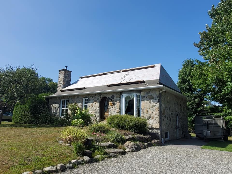 Maison en pierre avec toit gris, cheminée, bow-window, perron et allée sous un ciel bleu.