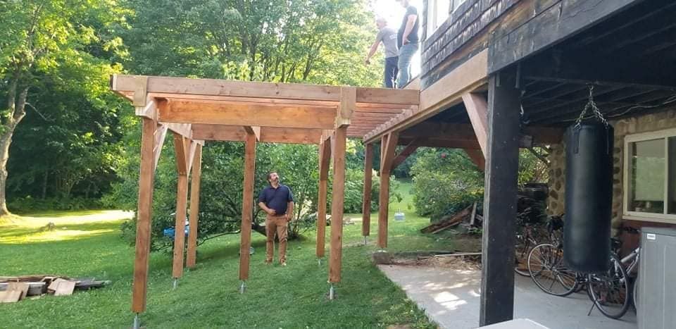 Des personnes construisent une pergola en bois attenante à une terrasse, dans un jardin. Un sac de frappe est suspendu