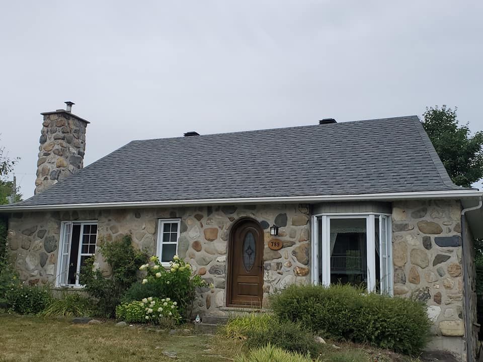 Maison en pierre au toit gris foncé, cheminée, fenêtres à encadrement blanc et porte en bois. Buissons verts et ciel couvert.