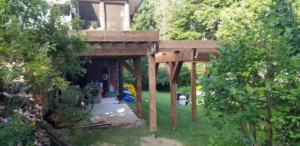Terrasse en bois prolongeant un bâtiment, soutenue par des poteaux, dans une cour verdoyante.