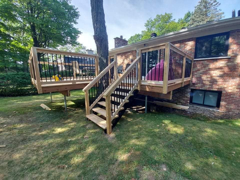 Terrasse en bois avec escalier attenant à une maison en briques dans un jardin gazonné, balustrade noire et grand arbre.