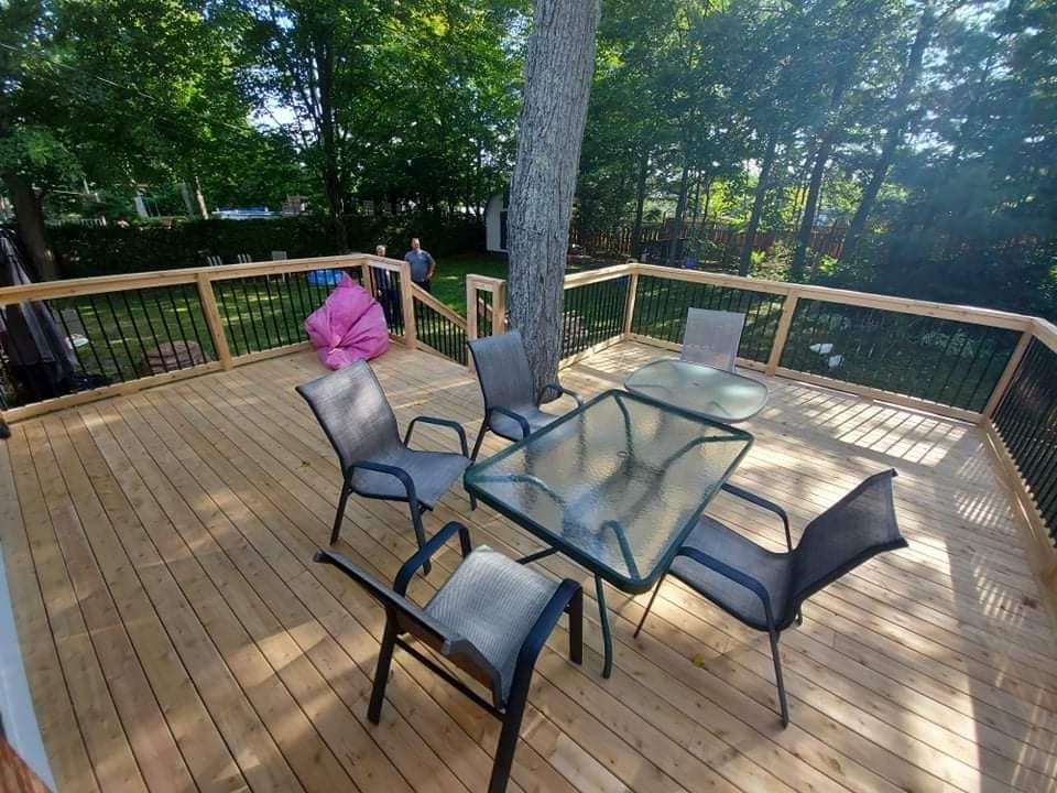 Terrasse en bois avec mobilier de jardin autour d'un arbre, entourée d'une balustrade noire, dans un jardin.