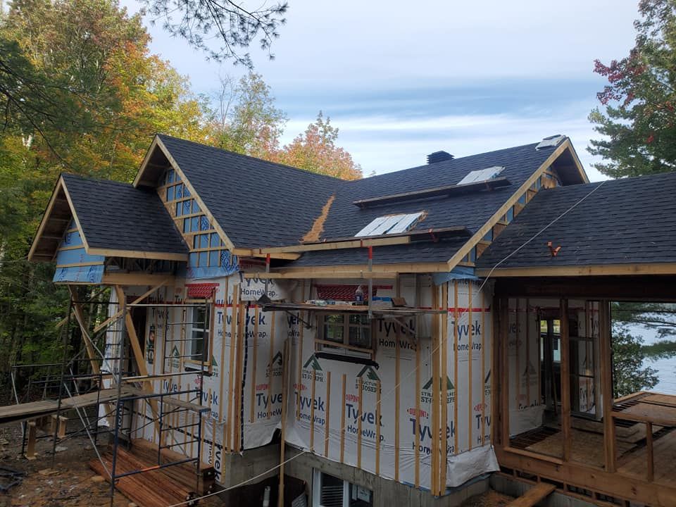 Maison en construction avec un toit en bardeaux foncés, une bâche bleue et des échafaudages, entourée d'arbres.