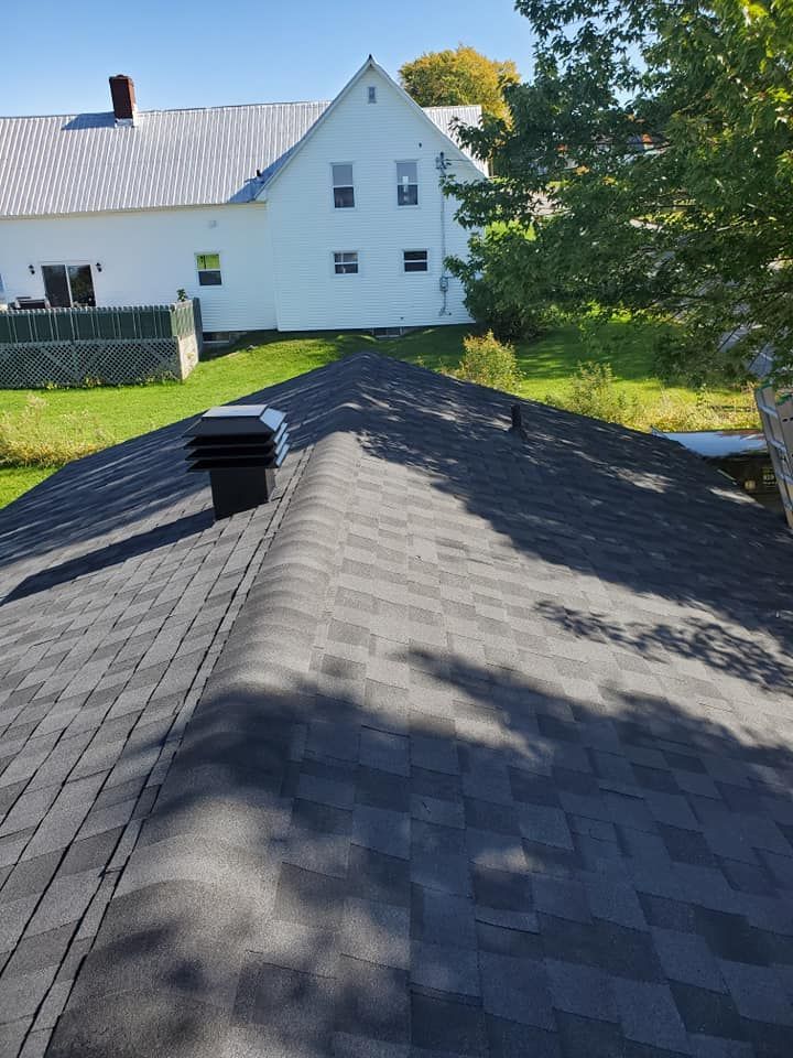 Toit en bardeaux foncés, grille d'aération noire, maison blanche en arrière-plan, herbe verte, ciel bleu.