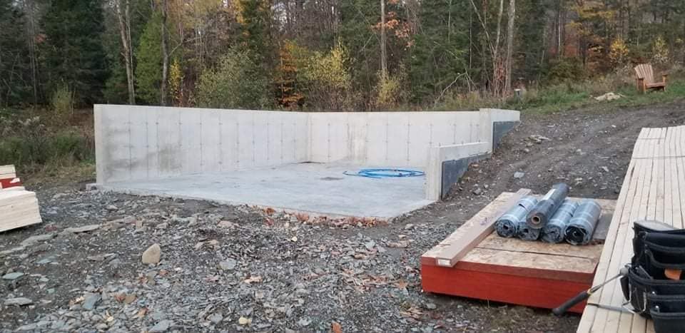Fondations en béton d'un bâtiment en construction, situé dans une zone boisée. Bois et matériaux de construction visibles.