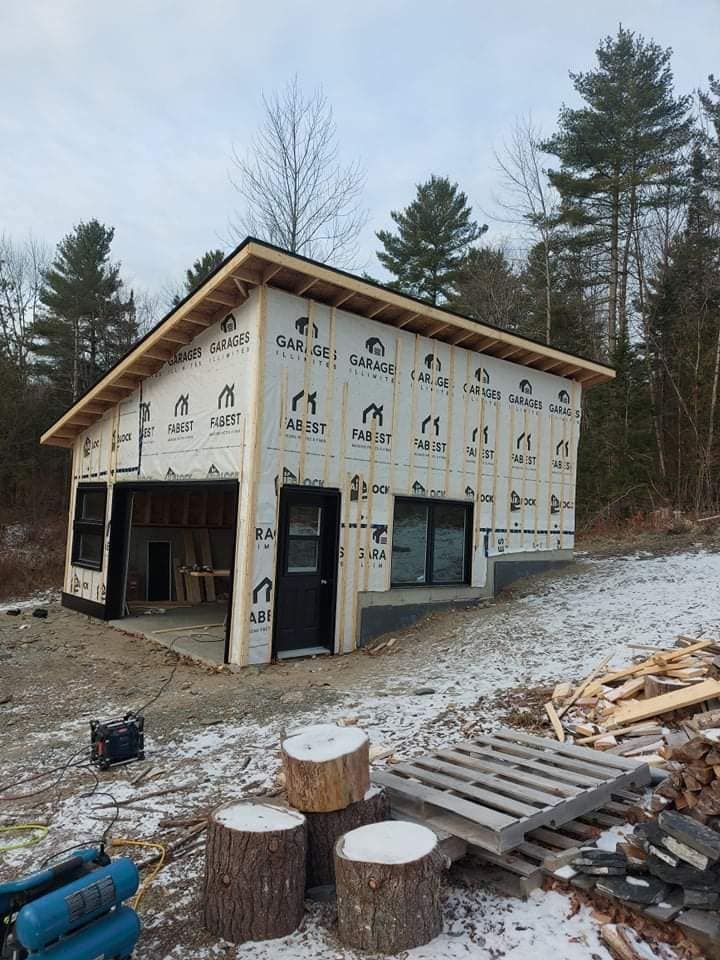 Un garage partiellement construit dans un décor enneigé, avec une porte noire, une ouverture de porte de garage