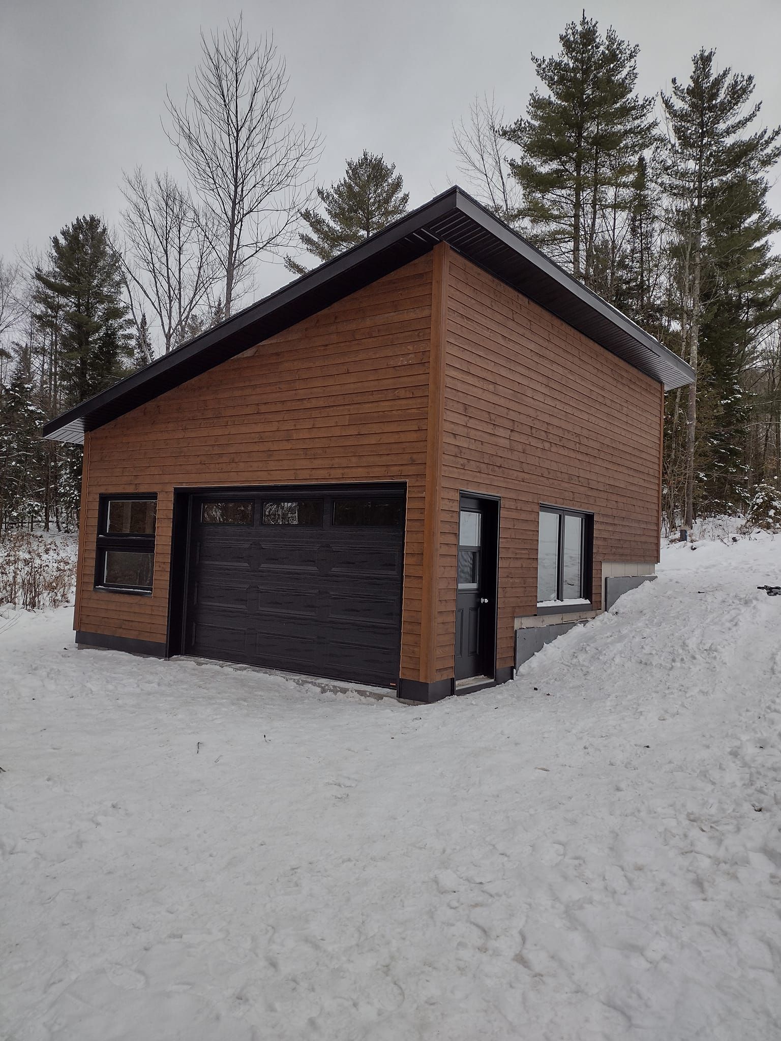 Garage en briques sous la neige, avec porte, fenêtres et toit noirs. Arbres en arrière-plan.