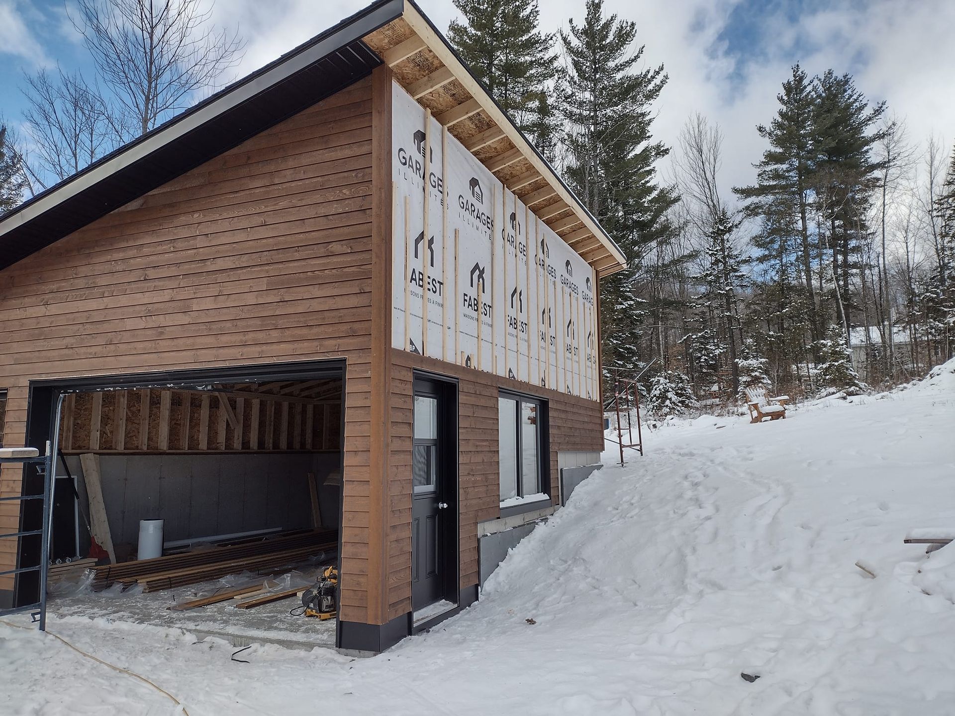 Garage en construction avec façade en briques, sol enneigé et arbres environnants.