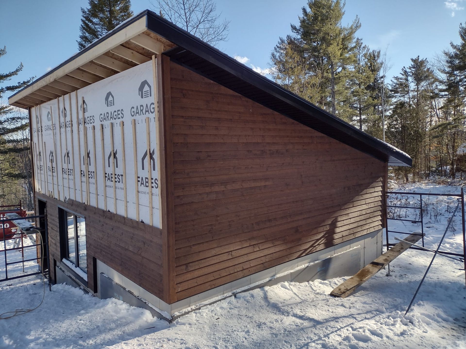 Extérieur du bâtiment avec bardage en bois, toit noir et neige au sol.