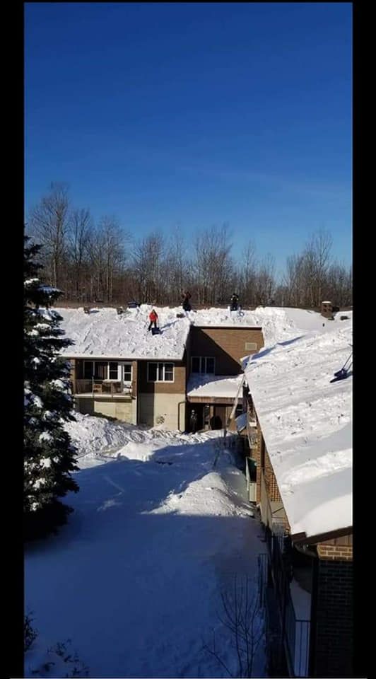 Maison enneigée avec une personne sur le toit, arbres et ciel bleu dégagé.