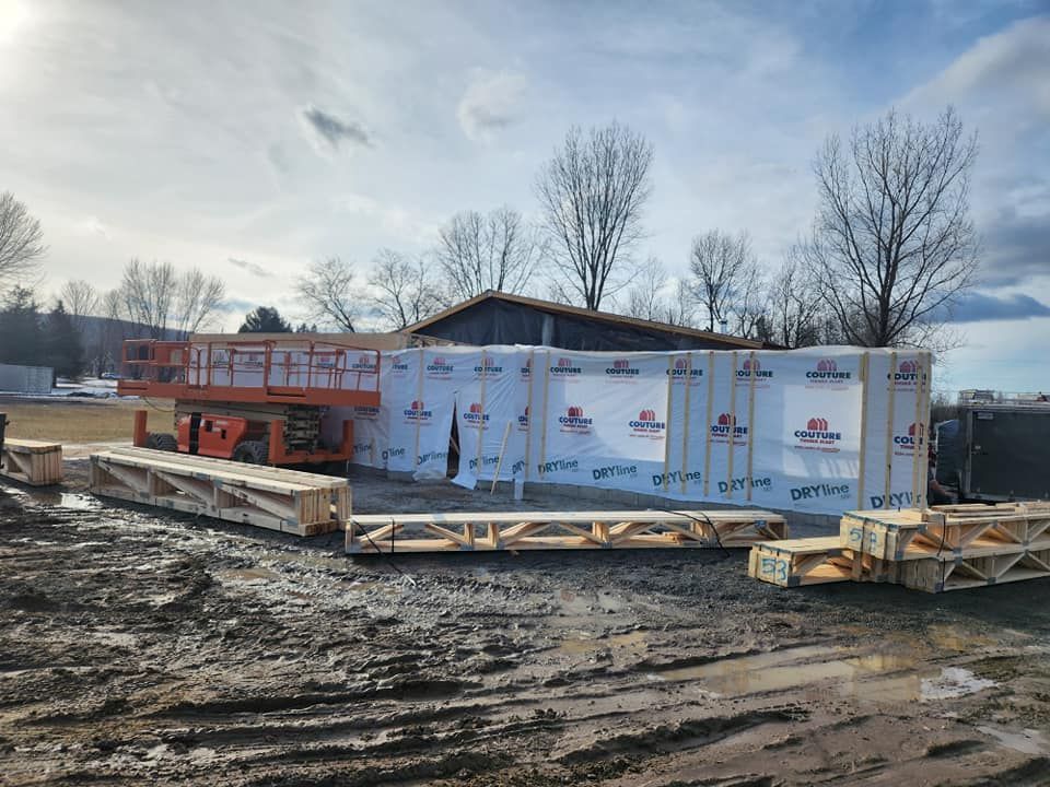 Chantier : Charpente en bois recouverte d'une bâche de protection ; nacelle élévatrice en arrière-plan ; sol boueux.