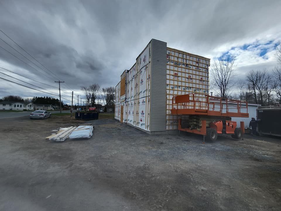 Façade du bâtiment en construction, isolation apparente. Un ascenseur est stationné à proximité. Ciel nuageux.
