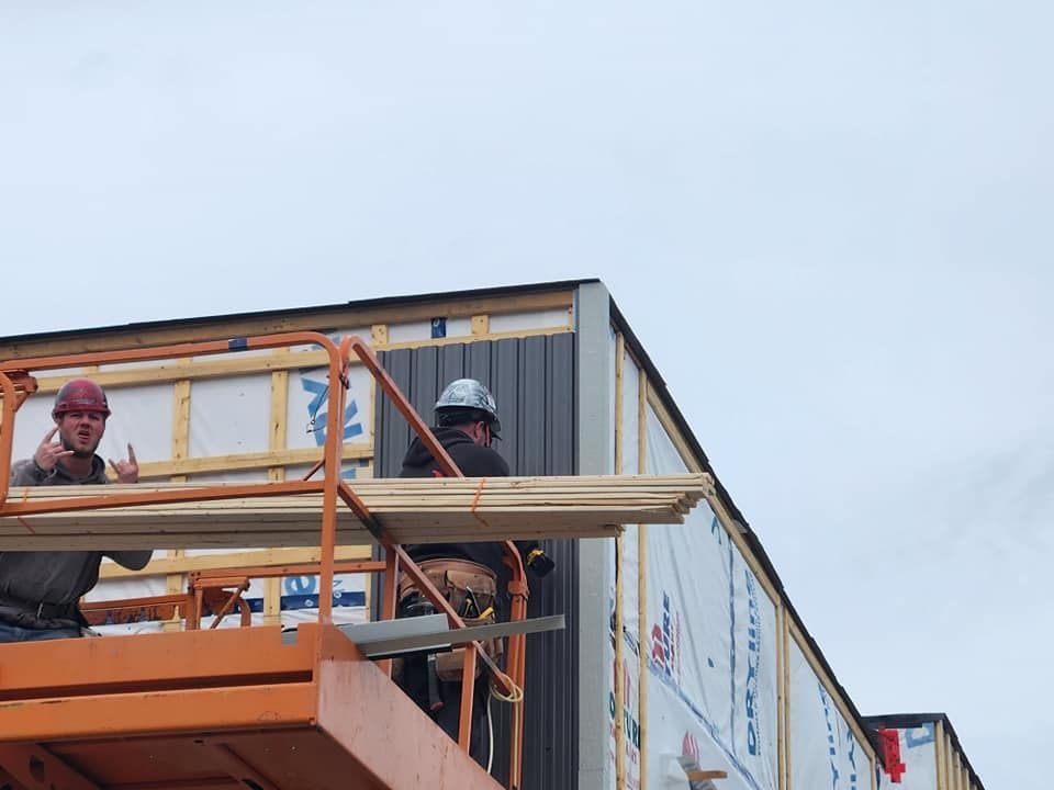 Des ouvriers du bâtiment installent un bardage sur un immeuble ; l'un d'eux fait un geste, ciel nuageux.