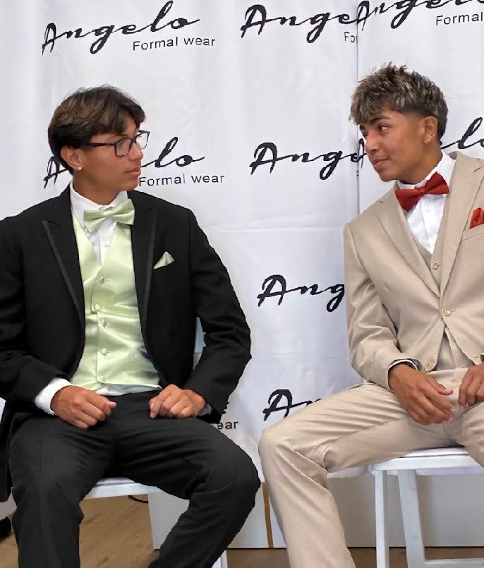 Two men in suits seated at a formalwear event backdrop, looking at each other.