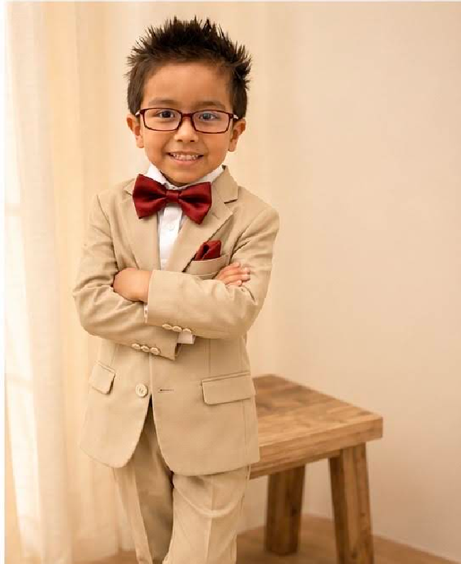 Child in a beige suit with red bow tie and glasses, smiling with arms crossed beside a wooden bench.