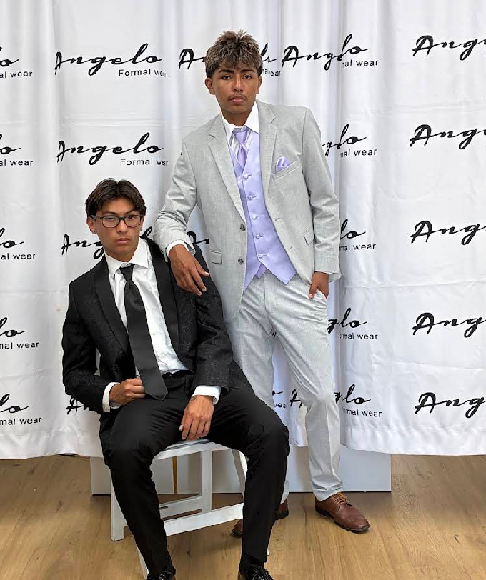 Two men in suits posing at an event backdrop, one seated and one standing.