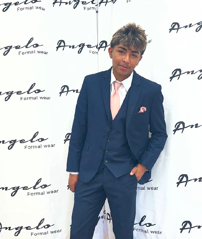 Person in a navy suit with pink tie and pocket square, posing at an Angelo event backdrop.