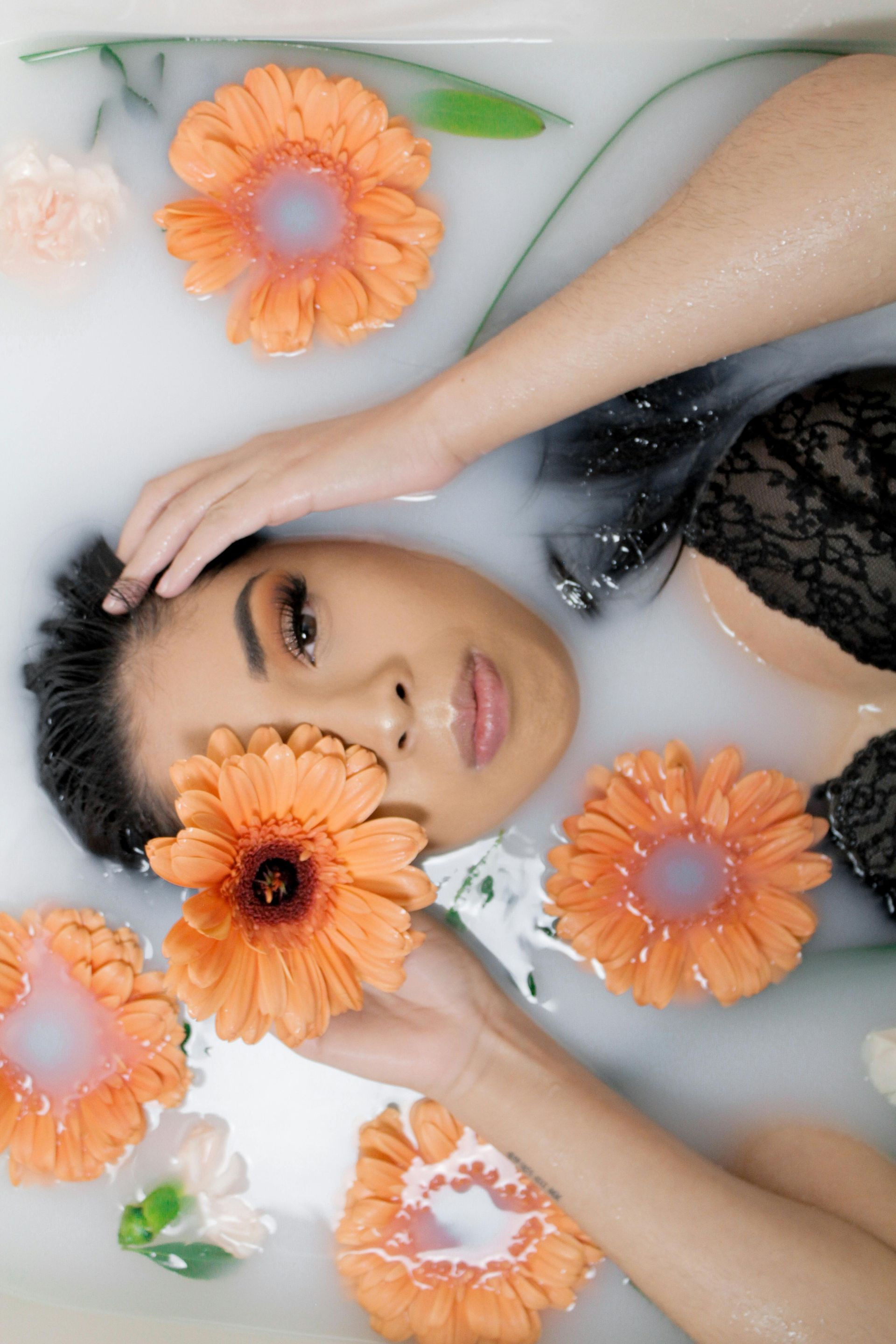 Person reclining in milky water, surrounded by orange flowers, with hands framing their face