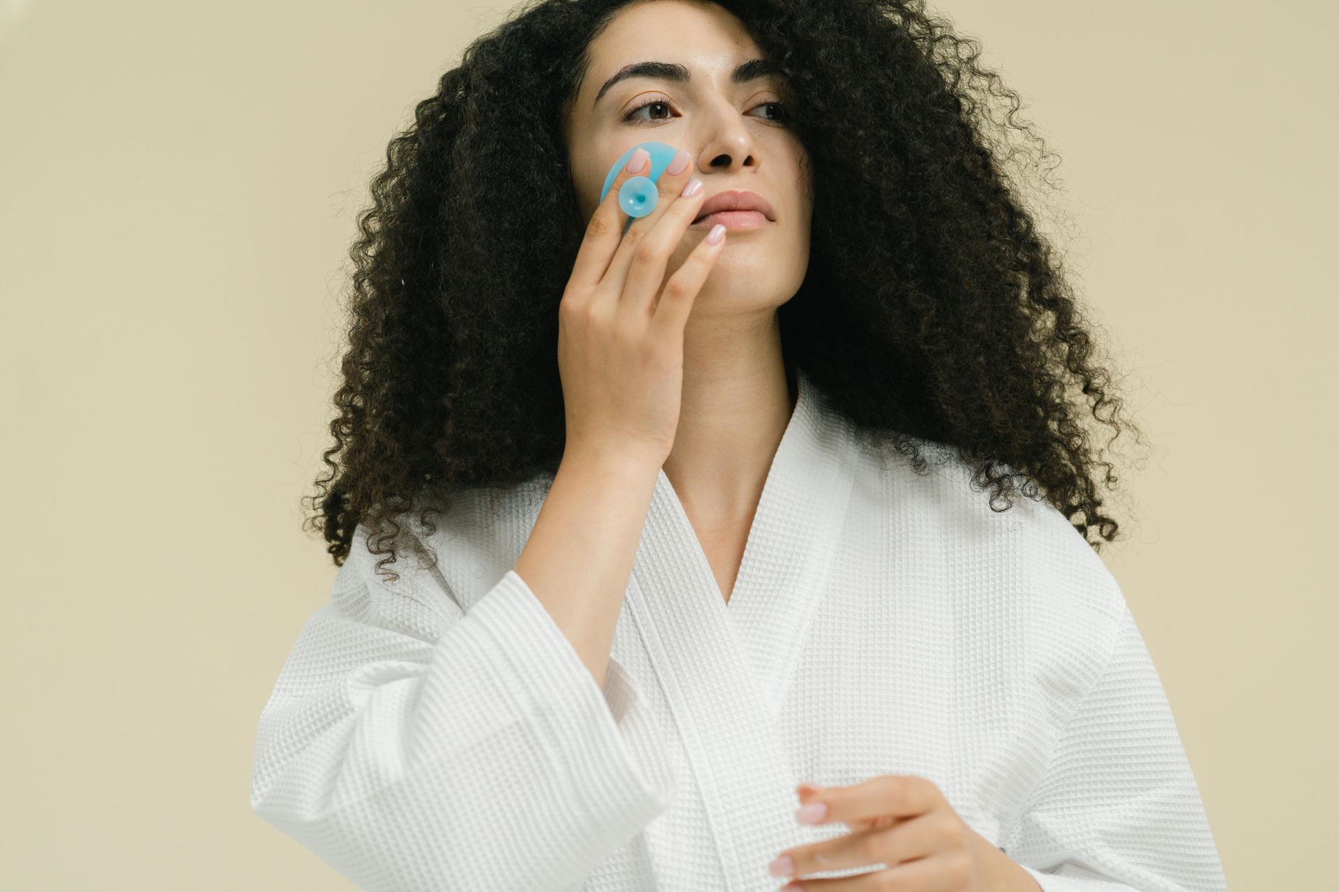 Woman in a white robe applying blue skincare roller to her cheek against a beige background