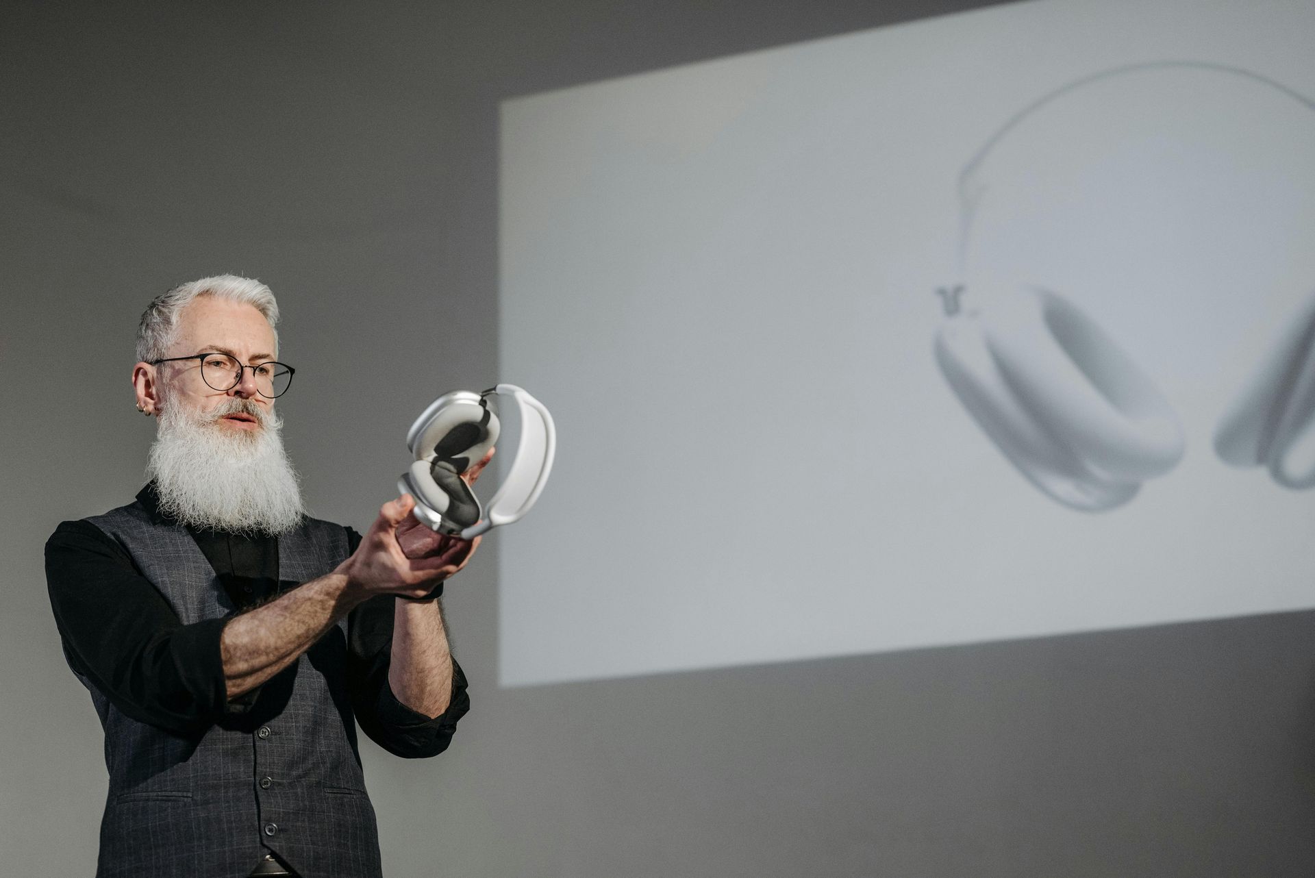 A man with a beard is holding a pair of headphones in front of a projection screen.