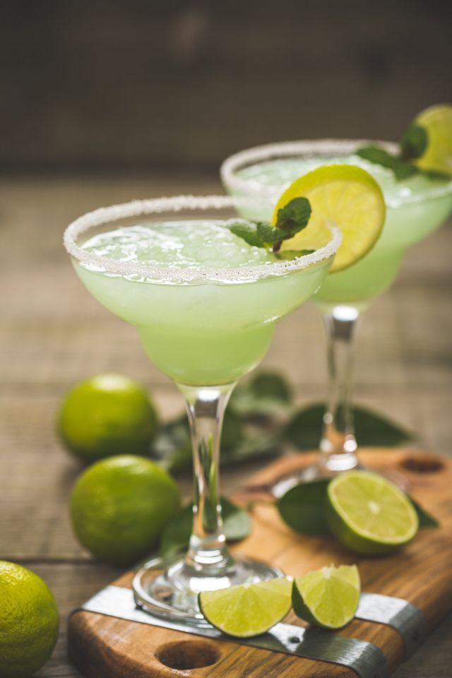 Two green margaritas with limes on a wooden cutting board.
