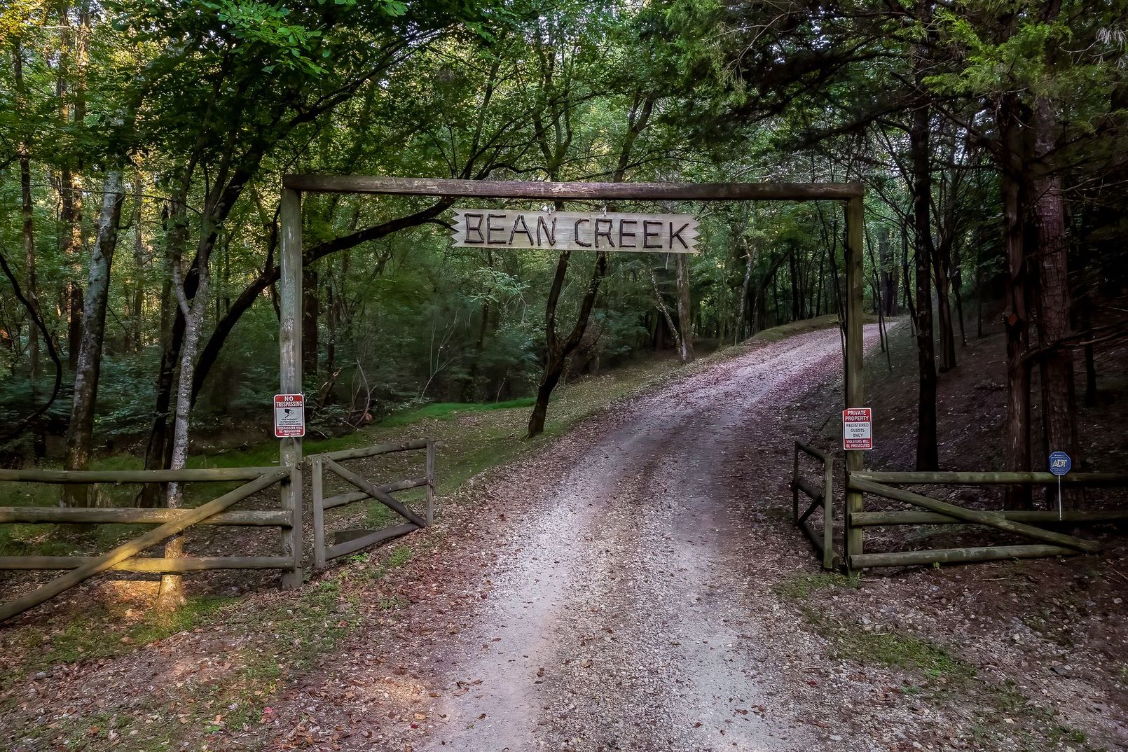 A dirt road enters a wooded area through a rustic wooden gate with a sign above that reads