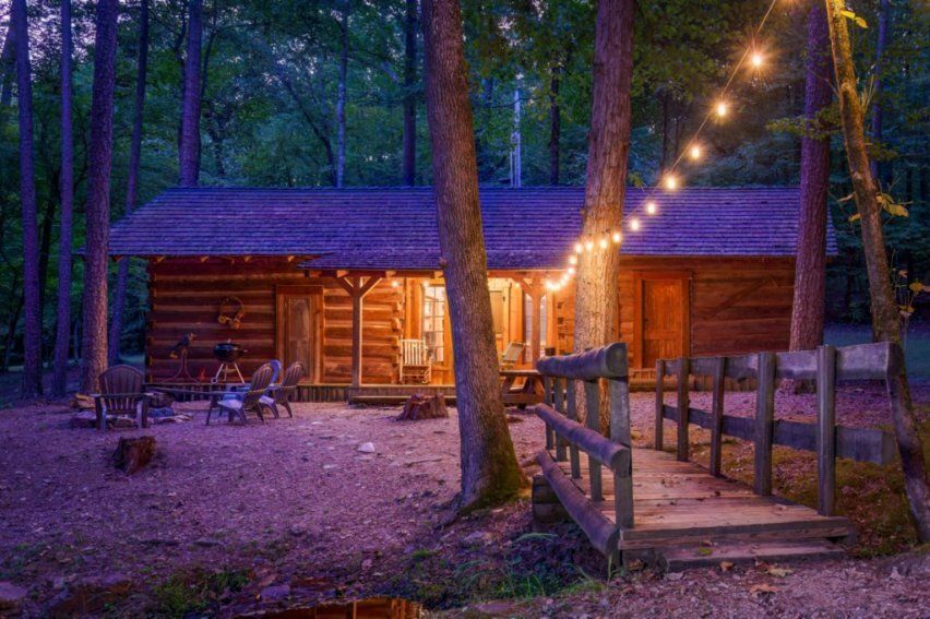 A rustic wooden log cabin in a forest at dusk, illuminated by glowing string lights and a small wooden footbridge.
