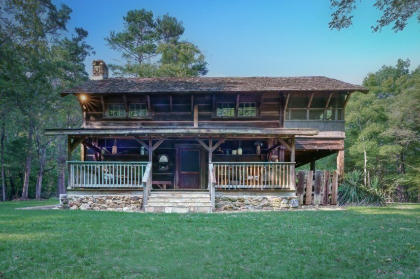 A rustic two-story log cabin with a stone foundation and a wide covered porch, nestled in a grassy clearing with trees.
