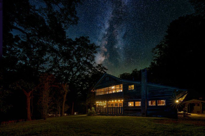 A two-story house illuminated with warm lights at night, set against a dark forest under a clear, starry Milky Way sky.